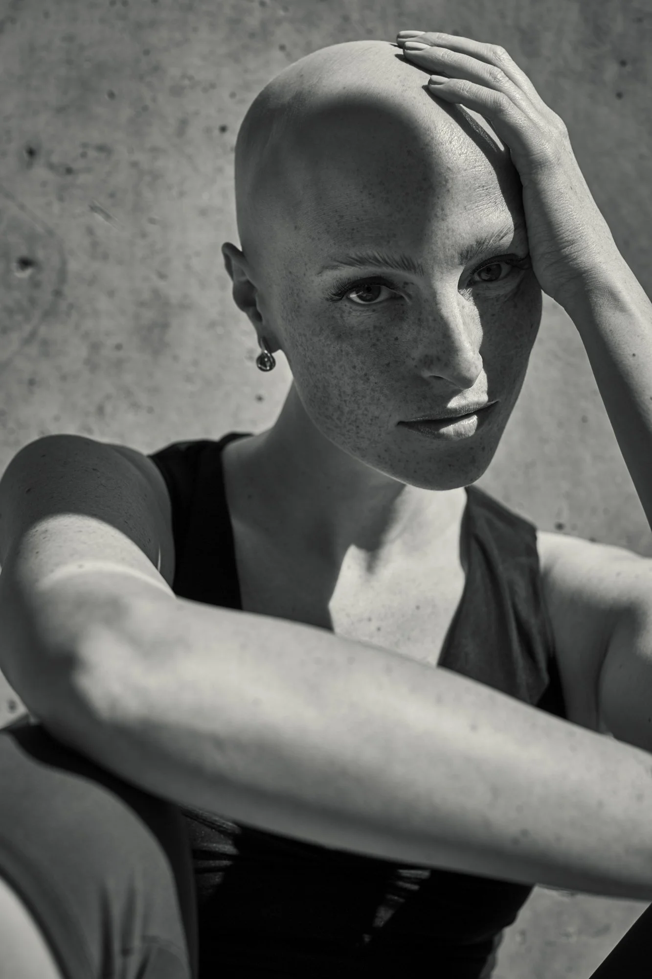 Black and white portrait of a woman with a shaved head, freckles, and earrings, looking directly at the camera with one hand on her head, taken outdoors in sunlight.