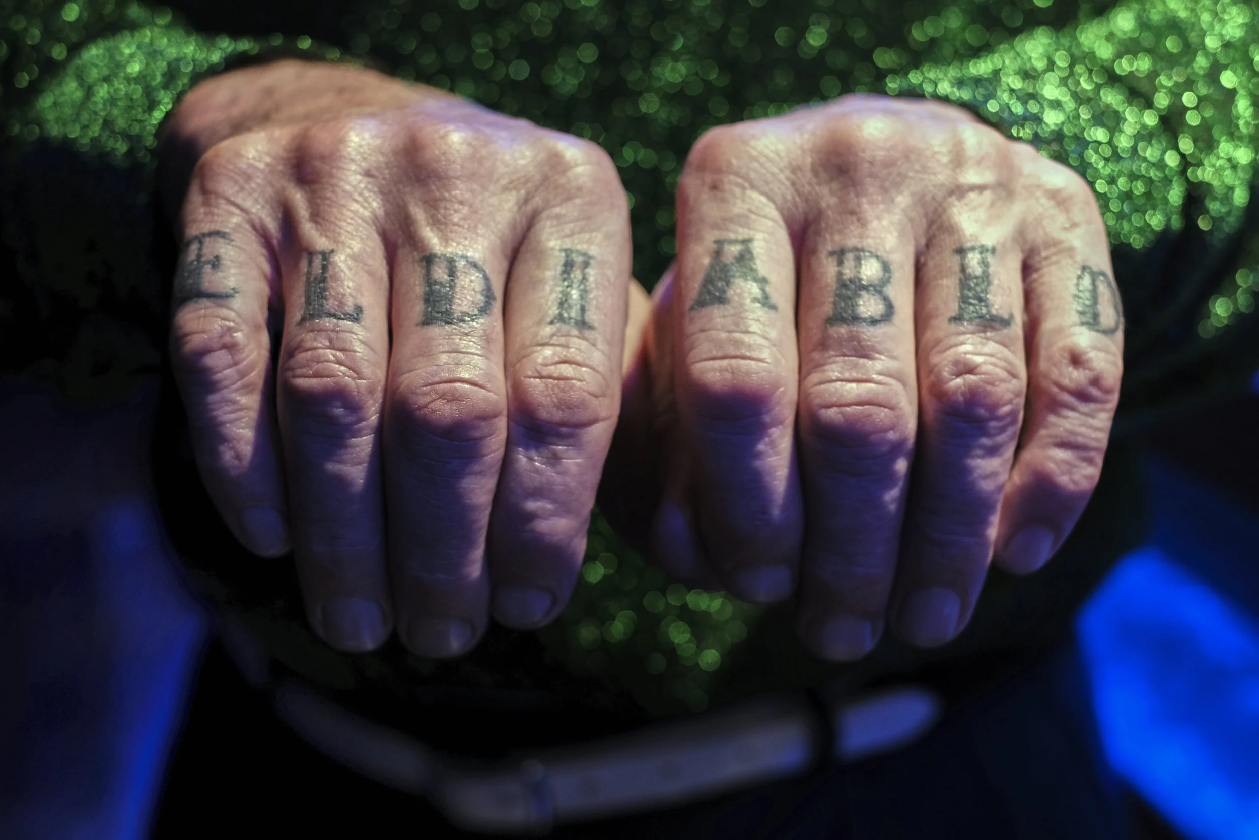 Close-up of two clenched fists with tattoos spelling 'SALDIA' on the fingers, against a sparkly green background.