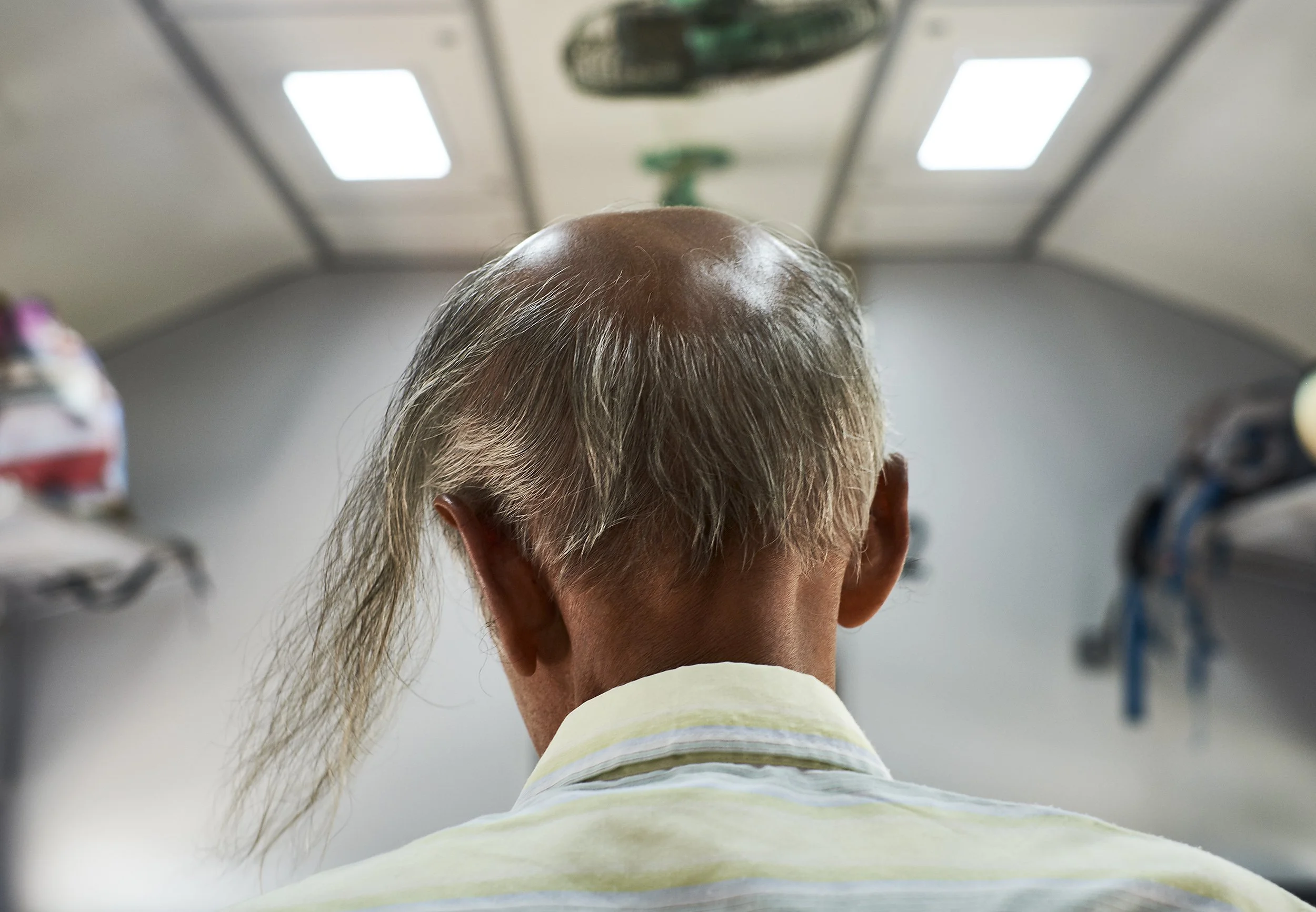 Back view of an elderly man with a bald head and long gray hair, wearing a striped shirt, inside a room with a ceiling light and shelves on the sides.