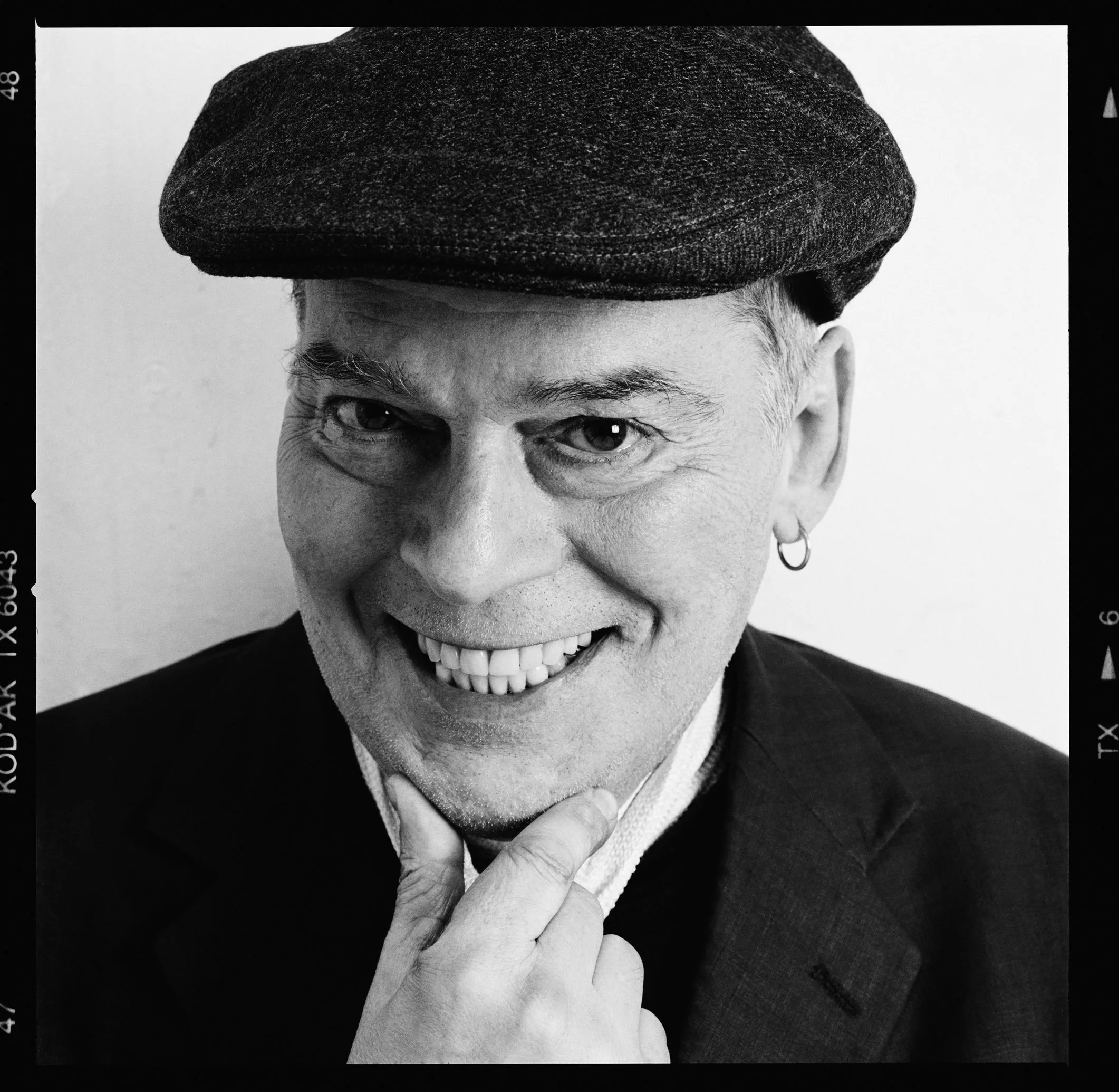 Black and white portrait of a smiling man wearing a beret, earring, and suit, with his hand resting on his chin.