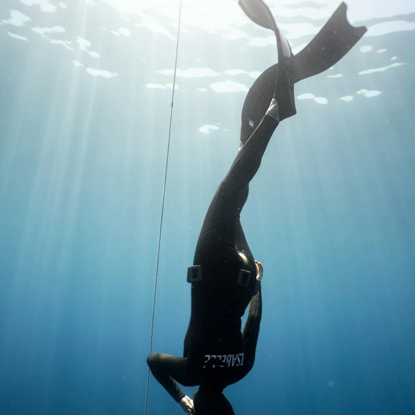A person freediving underwater, hanging upside down with fins and a wetsuit, with sunlight streaming through the water.