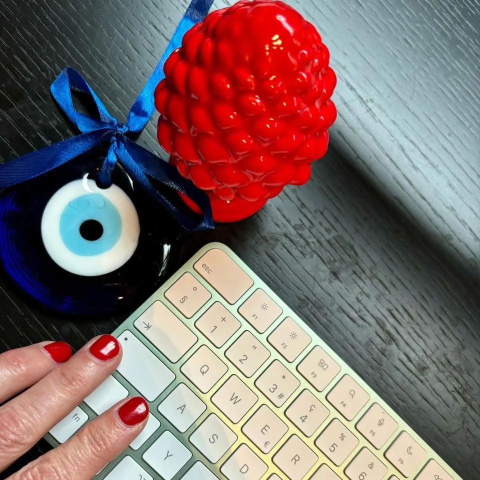 A glass apple-shaped container with a blue ribbon, an eye-shaped object, a red textured candle or decorative item, and a white keyboard with a person's hand with painted red nails resting on it, all on a dark wooden surface.