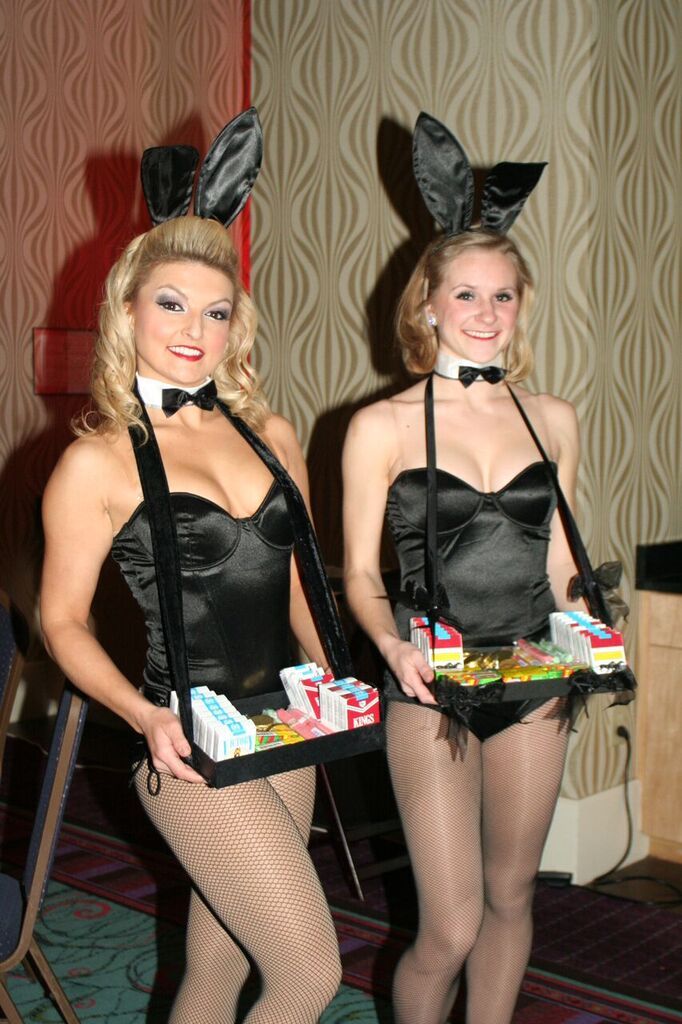 Two performers in black Playboy Bunny themed costumes holding cigarette trays to pass candy cigarettes and other party favors to guests at an event