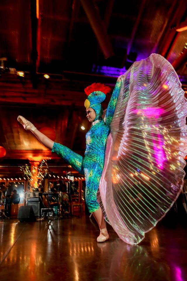 Bird Ballerina Performer dancing en pointe in a blue and green costume with iridescent wings and the face of a bird on her head