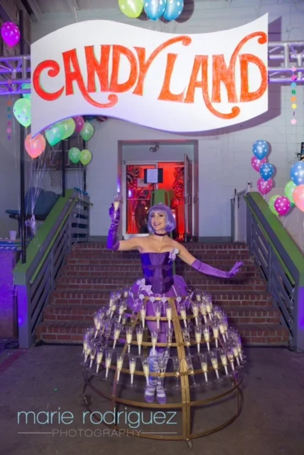 Champagne dress performer costumed in purple standing in from of a large sign reading "Candy Land"