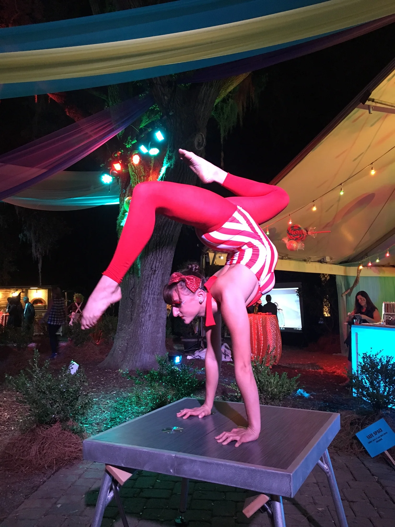 Hand balancing contortionist on top of a table costumed in red tights and a red and white striped leotard 
