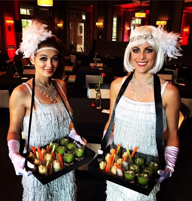 Two flapper performers in white fringe dresses, gloves, and feather headbands carrying cigarette trays filled with hors d'oeuvres to pass at a 1920s themed event