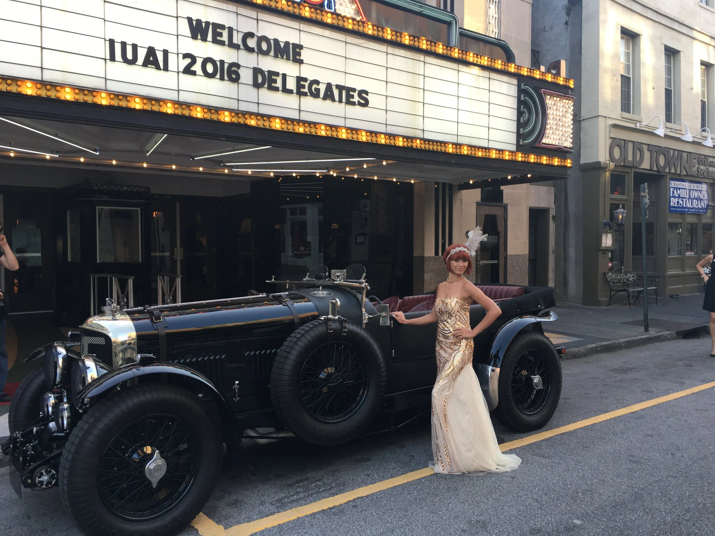 Performer in a gold art deco gown and feather headpiece standing beside a vintage car for a 1920s themed event in Charleston, SC