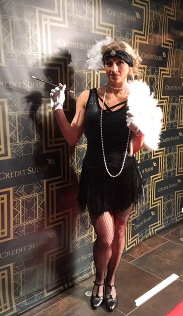 Flapper performer costumes in a black fringe dress, white feather boa, white gloves, and black and white feather headband holding a long cigarette holder