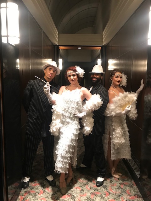 Two female flapper performers costumed in a white fringe dress, feather boa, feather headband, and long cigarette holder and two male performers costumed in black pin stripe suits and white fedoras