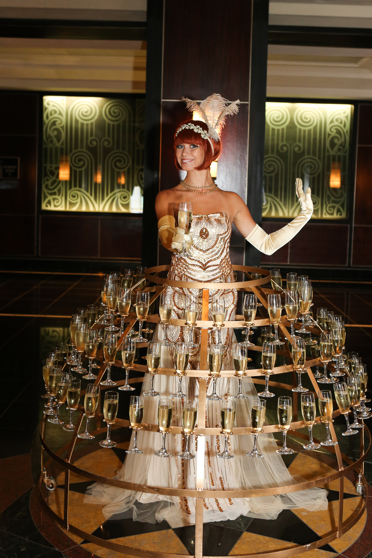 Champagne dress performer costumed in a gold art deco gown, feather headpiece, and gloves handing a glass of champagne forward at a 1920s themed event in Charleston, SC