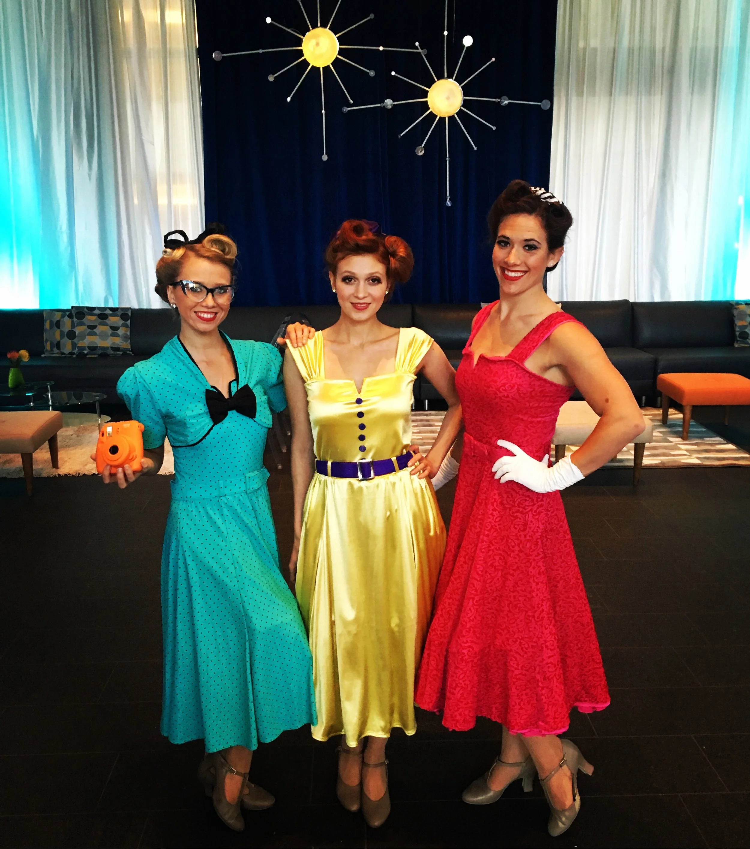 Three performers each in a red, yellow, or blue dress posing for a photo at a retro themed event