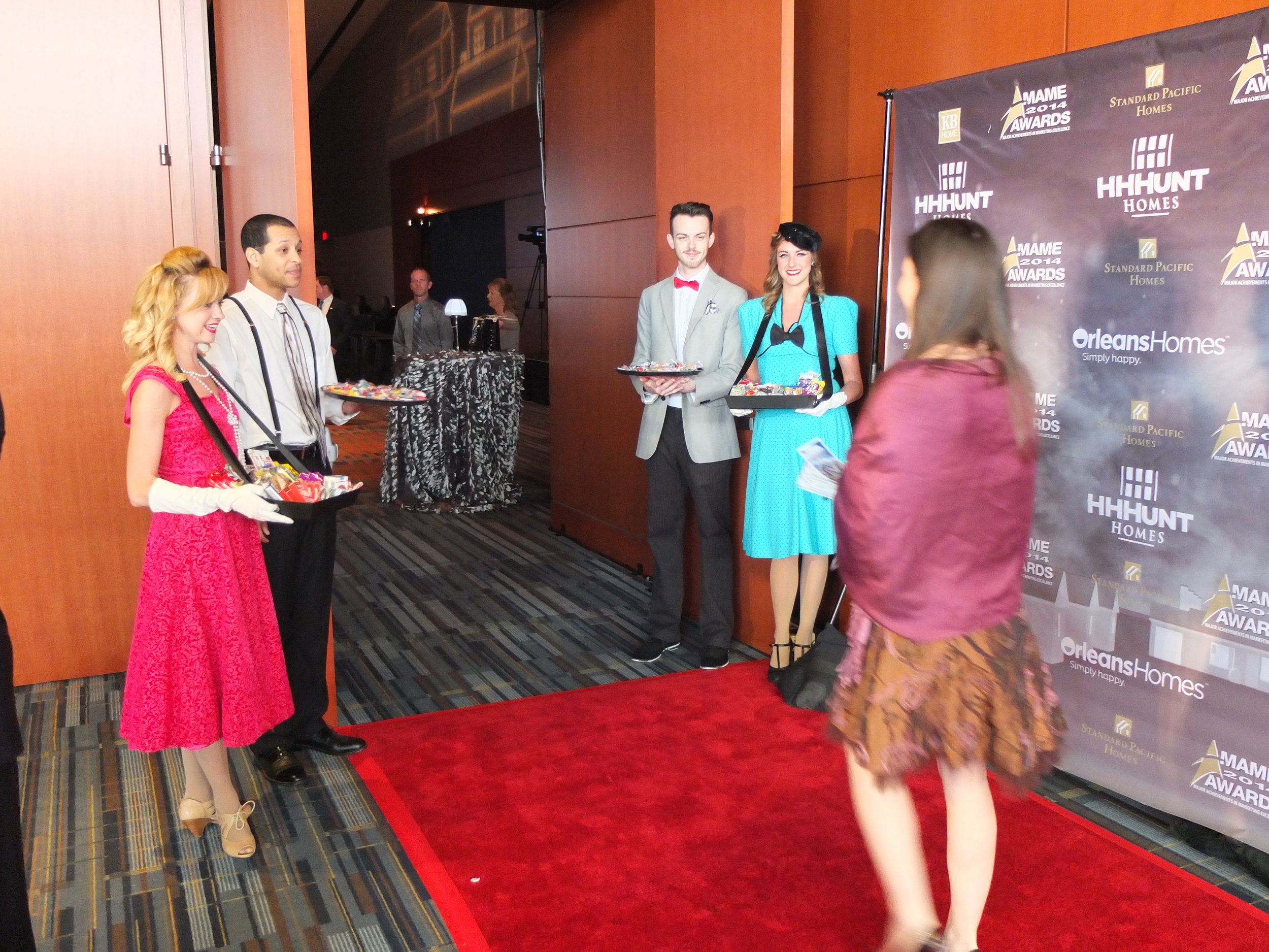 Four performers costumed in retro dresses and suits carrying cigarette trays to welcome guests at the end of a red carpet with an hors d'oeuvres on their way into an event