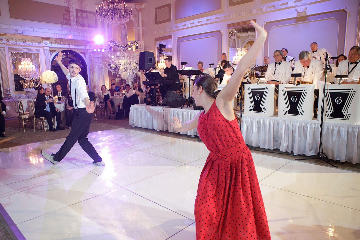 Male dancer in slacks, a white button down, and suspenders and a female dancer in a red dress performing a duet on the dance floor in front of a retro style big band