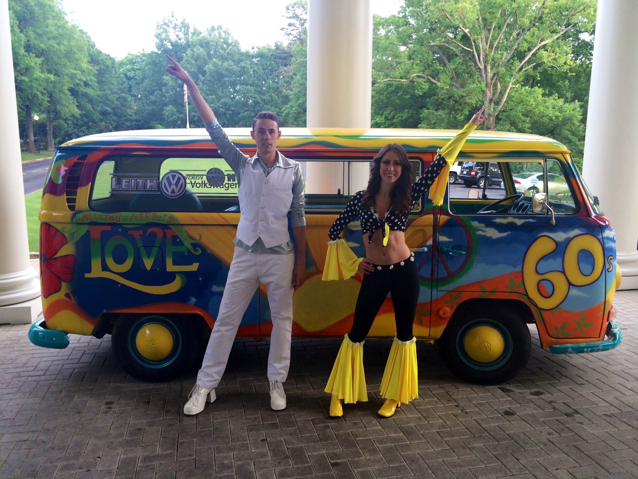 A female performer in a yellow and black floral bell-bottomed pant and bell-bottom sleeve crop top and a male performer costumed in white and silver posed in front of a 1960s hippie van