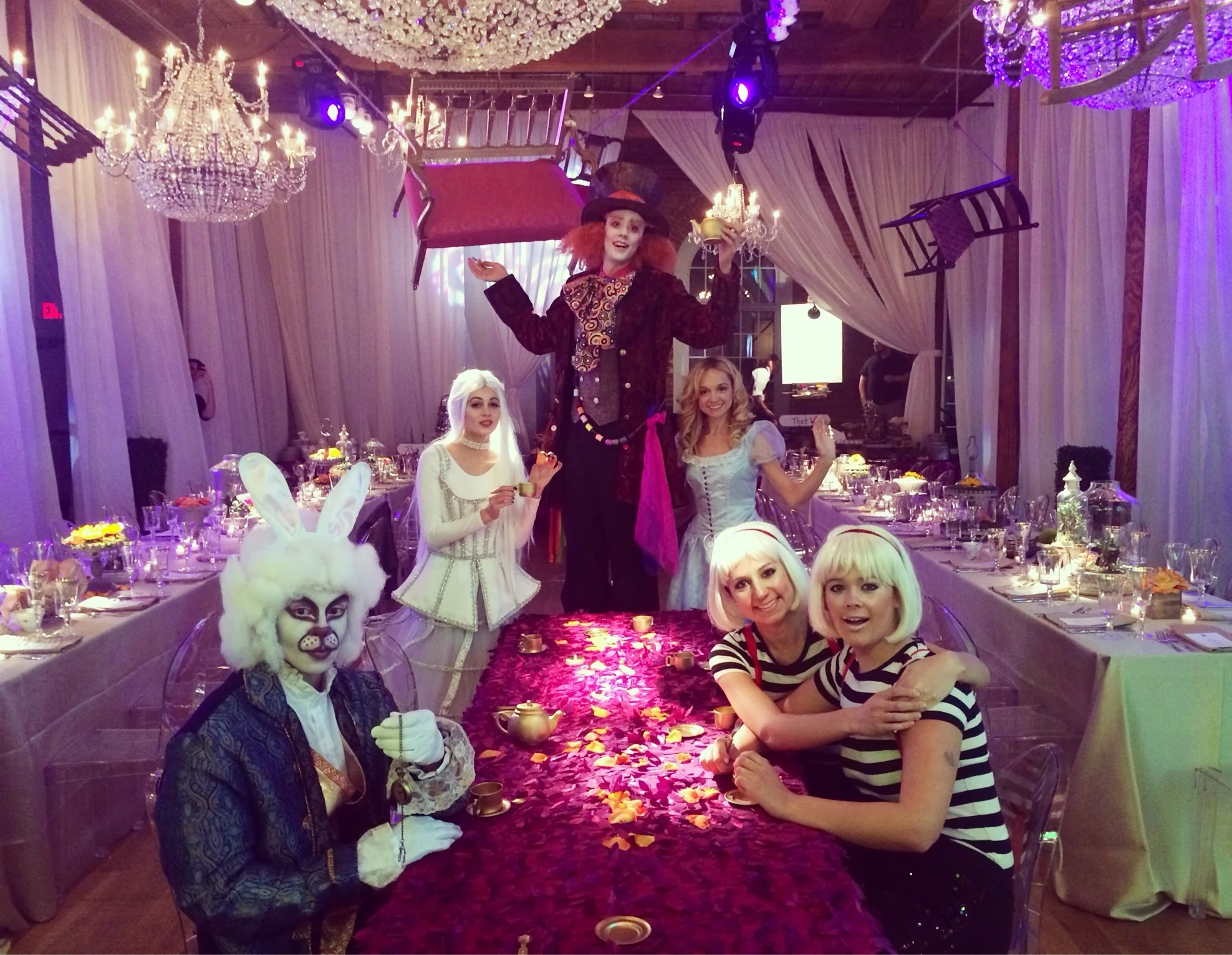 Six performers in White Rabbit, White Queen, Alice, Mad Hatter, and Tweedle Dees inspired costumes for an Alice in Wonderland themed party