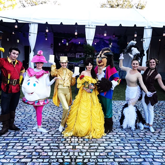 Seven performers in costume as Belle, the Beast, Lumiere, Mrs. Potts, Gaston, Feather Duster, and Cogsworth inspired by characters posing for a photo at a Beauty and the Beast themed event