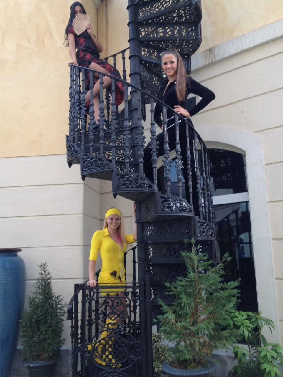 Three performers costumed in either yellow, black, or red and black posed on a spiral staircase