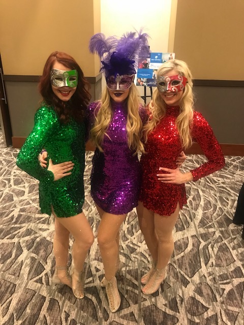 Three Mardi Gras performers costumed in sequined dresses and masks in either green, purple, or red