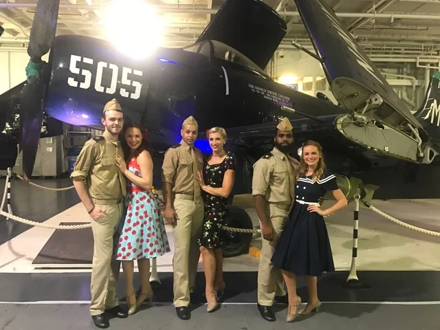 Three male performers costumed in World War II military uniforms and three female performers in 1950s dresses posed in front of a retro prop plane