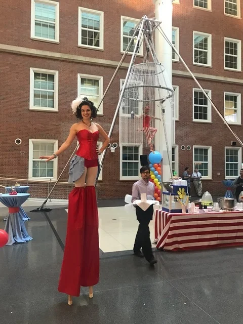Female stilt walker costumed in red with a grey bustle skirt, posed in front of an aerial quad rig with an aerialist in a candy cane inspired costume suspended inside an aerial chandelier on trapeze