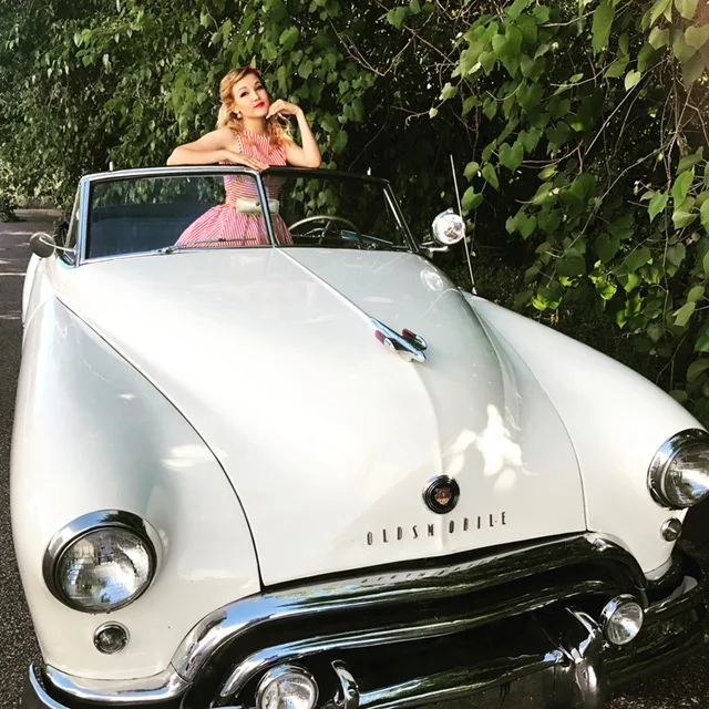 Performer costumed in a red and white striped dress posing for a photo in a retro car