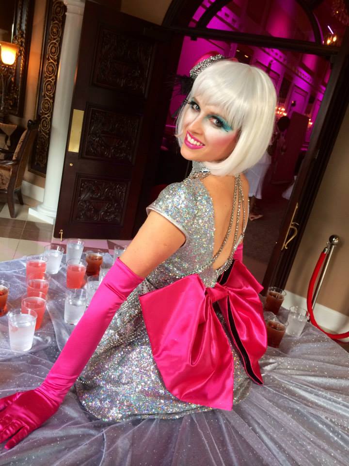 Performer costumed in silver with a big pink bow and pink gloves, living inside a large round table serving specialty drinks for guests at an event
