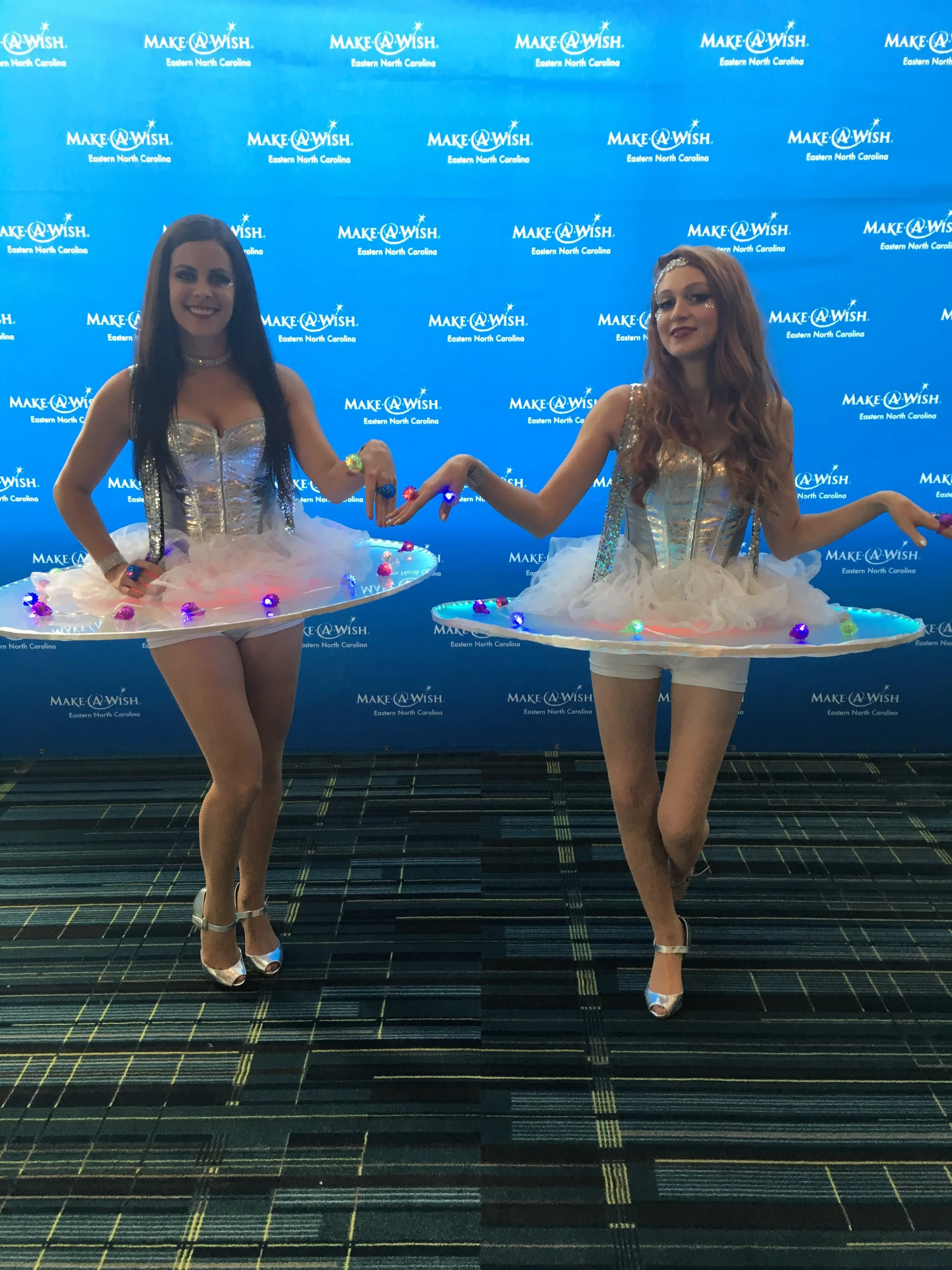 Two performers with an attached table around their waists to pass light up ring party favors and costumed in silver