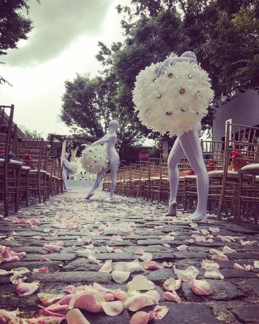 Two Flower Ball performers in white costumes that cover full body and face, with two large balls covered in white flowers posed outside on cobble stones for an event