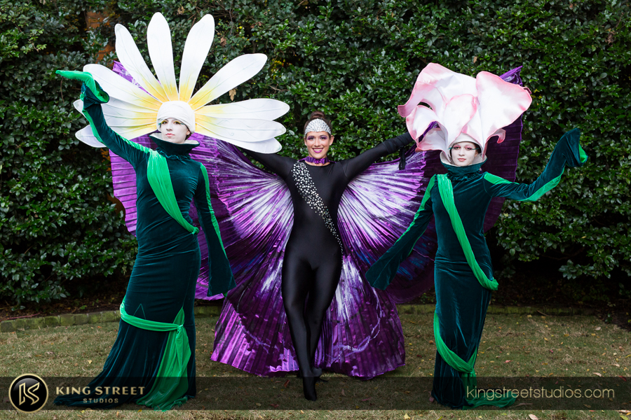 Two performers costumed in a large white flower headpiece with yellow or pink tips and a green vine-like gown. One performer costumed in black with large purple butterfly wings.