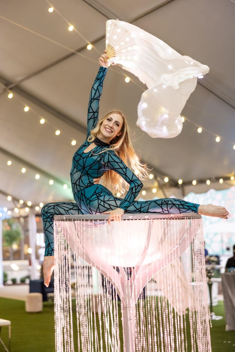Performer in a teal costume with a long white fan in a stag position on top of a huge martini glass covered in clear beads