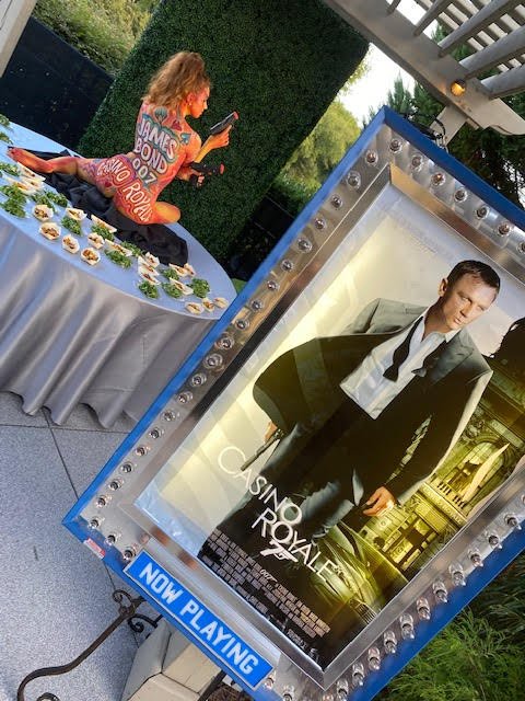 1967 Casino Royal inspired body painted performer positioned nearby a movie poster featuring Daniel Craig at a James Bond themed event