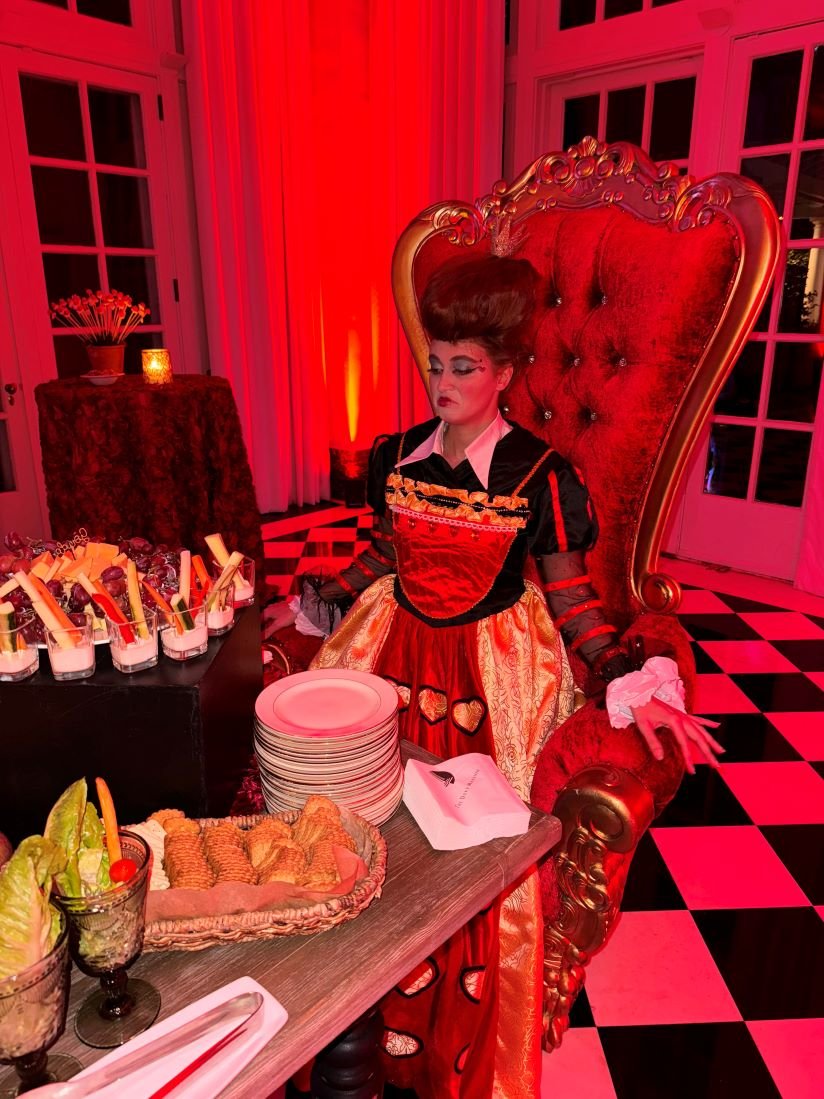 A performer in a Red Queen inspired costume for an Alice in Wonderland themed party sneering over a table filled with a buffet of food