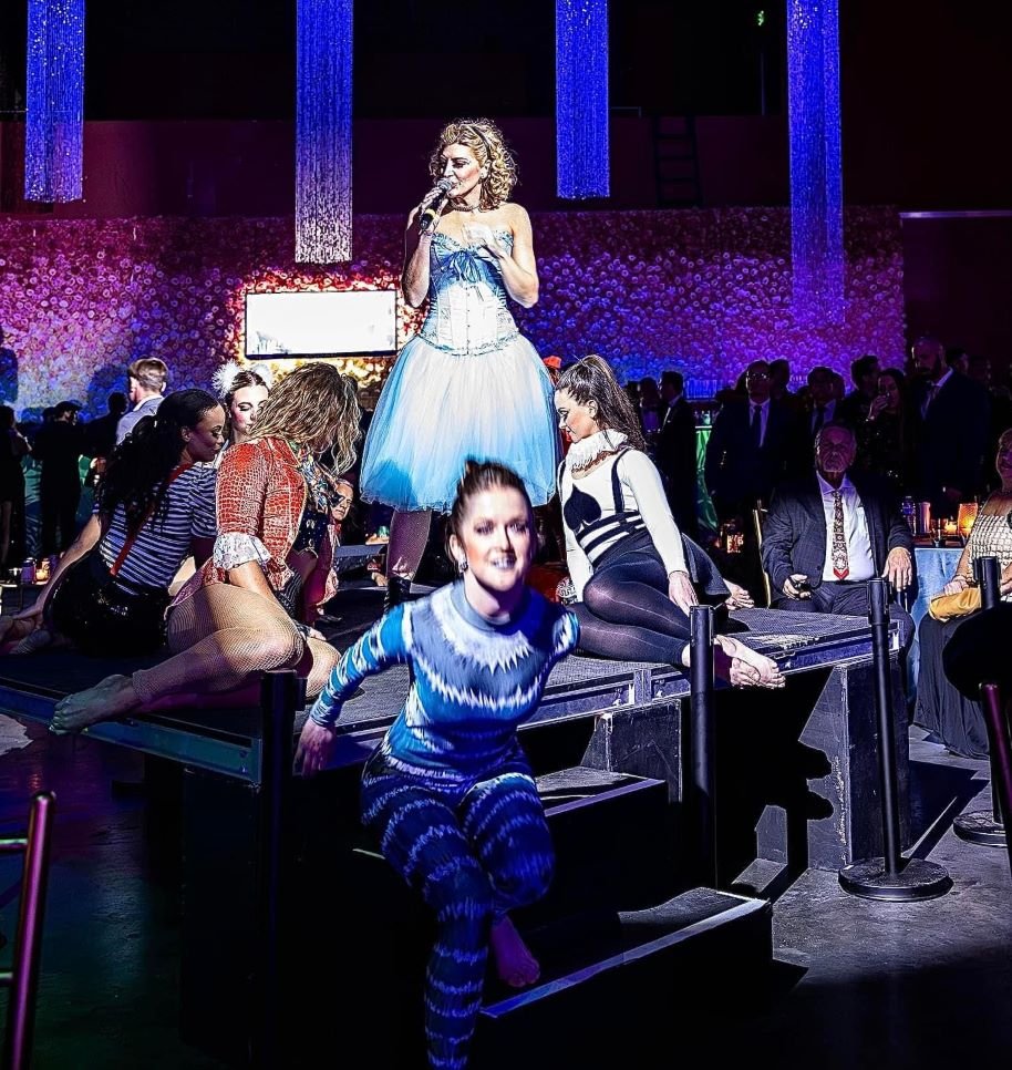 Four dancers in Tweedle Dee, Mad Hatter, blue Cheshire Cat, and Playing Card inspired costumes sitting around a singer in an Alice inspired costume performing for an Alice in Wonderland themed party