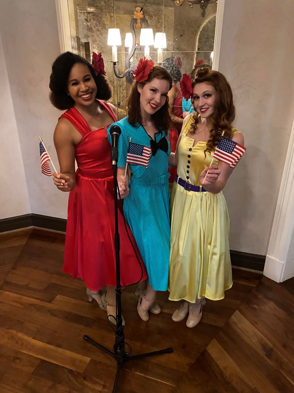 Three performers each in a red, yellow, or blue dress holding small American flags posing for a photo at a retro themed event