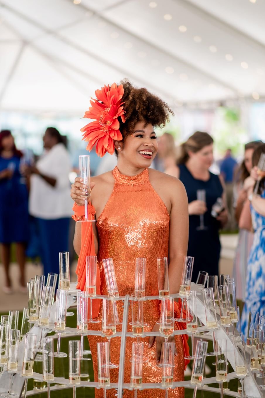 Performer in a metal champagne skirt, her skirt made of champagne flutes, costumed in an orange gown with orange flower in her hair at an outdoor event under a white tent