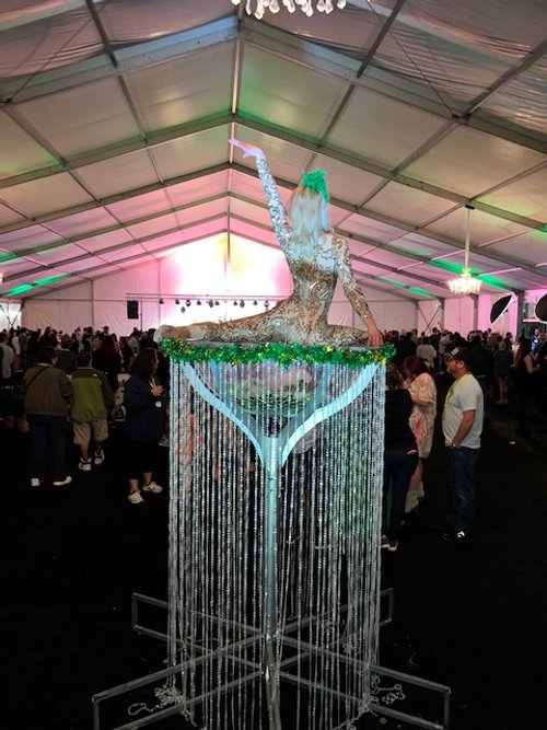 Performer in a gold costume and green feather headpiece in a double stag position on top of a huge martini glass covered in clear beads