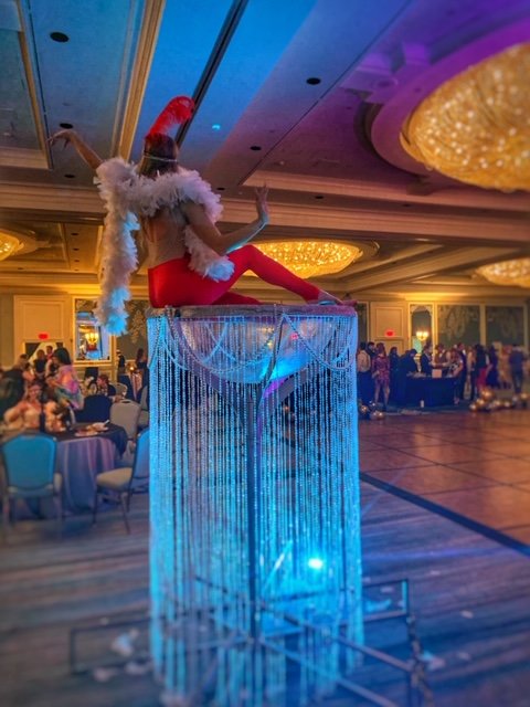 Performer in a red costume with a white feather boa sitting on top of a huge martini glass covered in clear beads