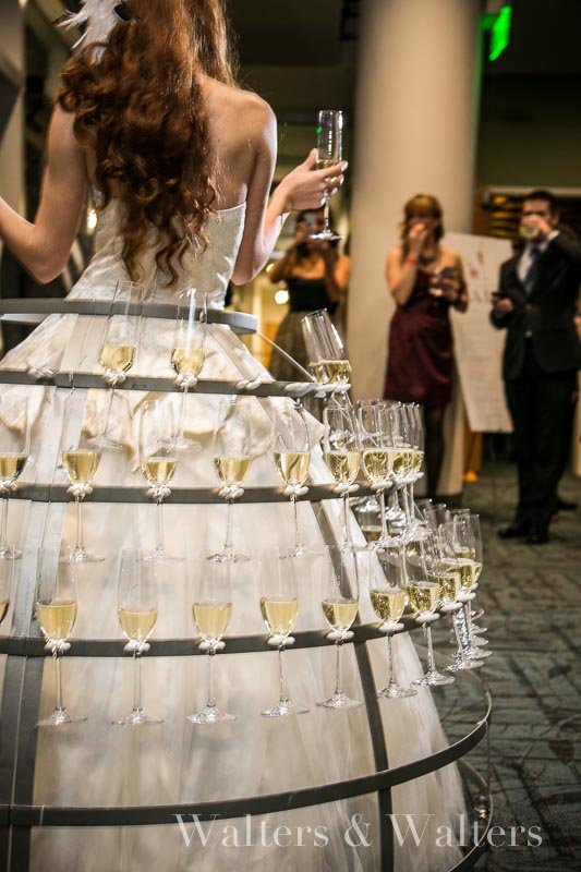 Performer in a metal champagne skirt, her skirt made of champagne flutes, costumed in a white gown handing one of the flutes to a guest at an event