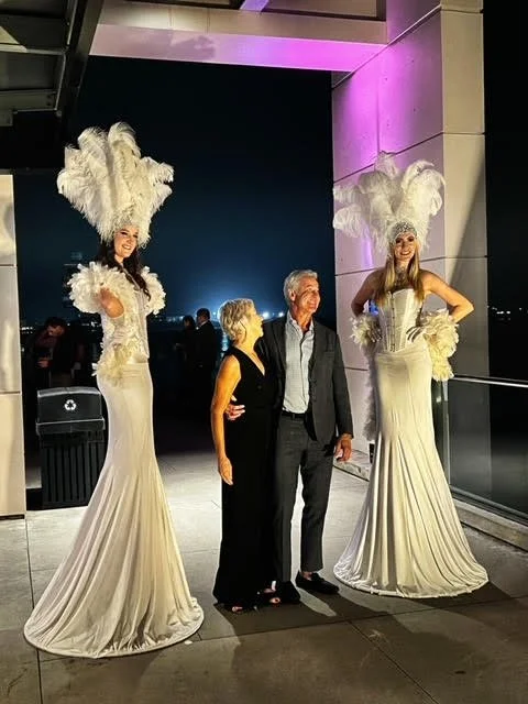 Two grand entrance performers much taller than any guest in white costumes with feather headpieces, feather boas, corsets, and long skirts