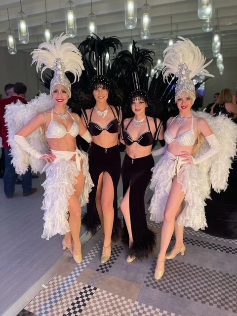 Four showgirl performers in either white or black costume bikini top, long skirt, feather headpiece, and feather backpack