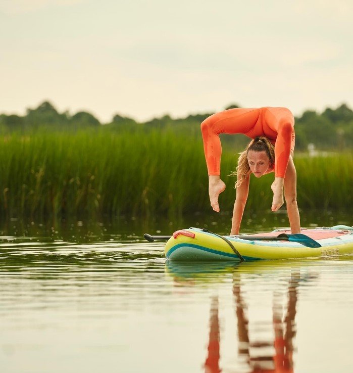 Contortionist in an orange costume performing a handstand on top of a Stand Up Paddle Board floating on a creek in Charleston, South Carolina