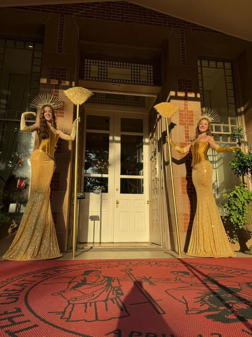 Two grand entrance performers much taller than any guest in gold costumes with gold headpieces, gloves, corsets, and long sequin skirts