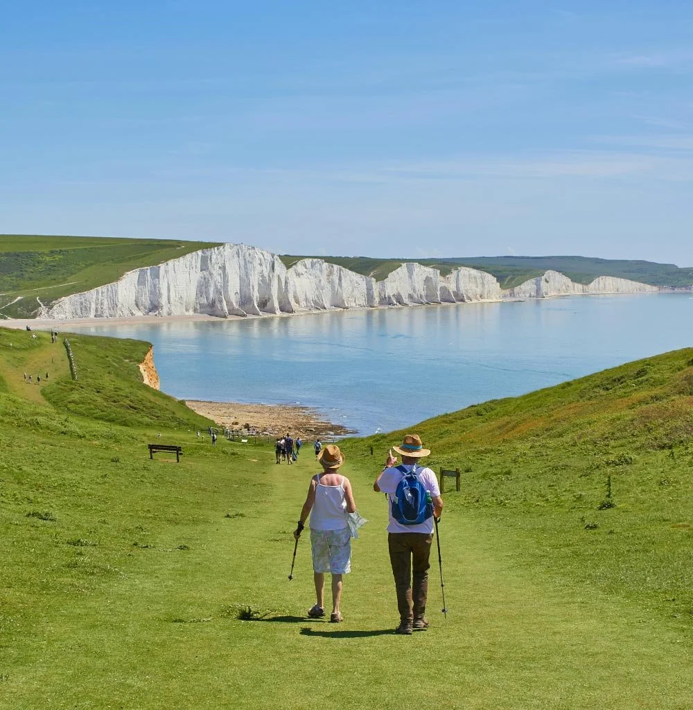 Retired couple walking along a grassy trail toward white chalk cliffs by the sea on a sunny day.