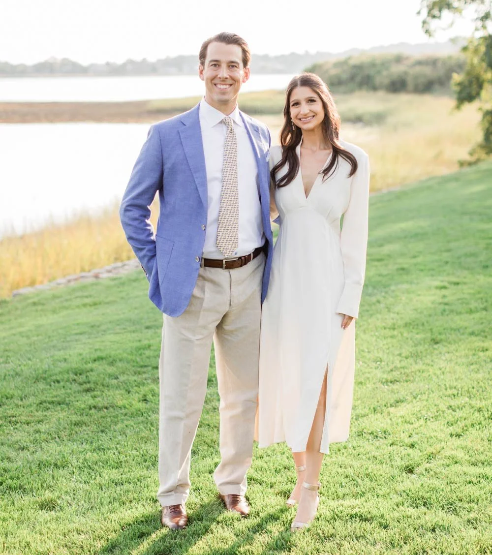 A smiling man and woman standing outdoors on grass near water, dressed in semi-formal attire.