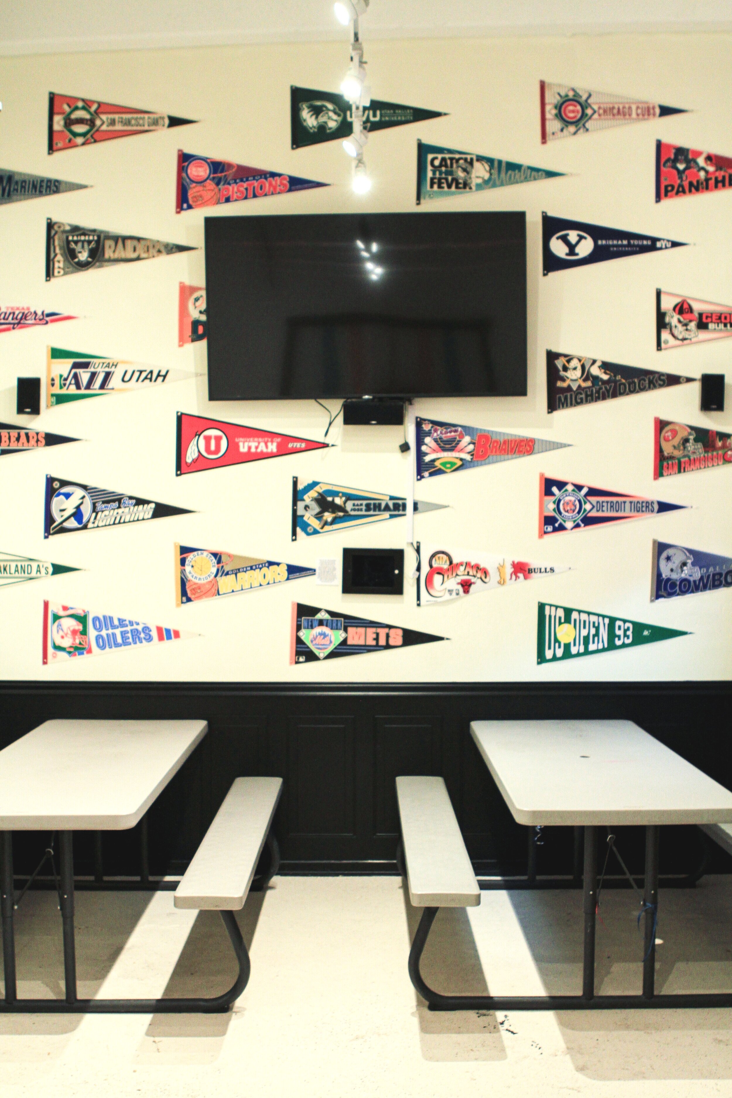  two picnic tables below a flat screen tv on a wall of sport and college pennants 