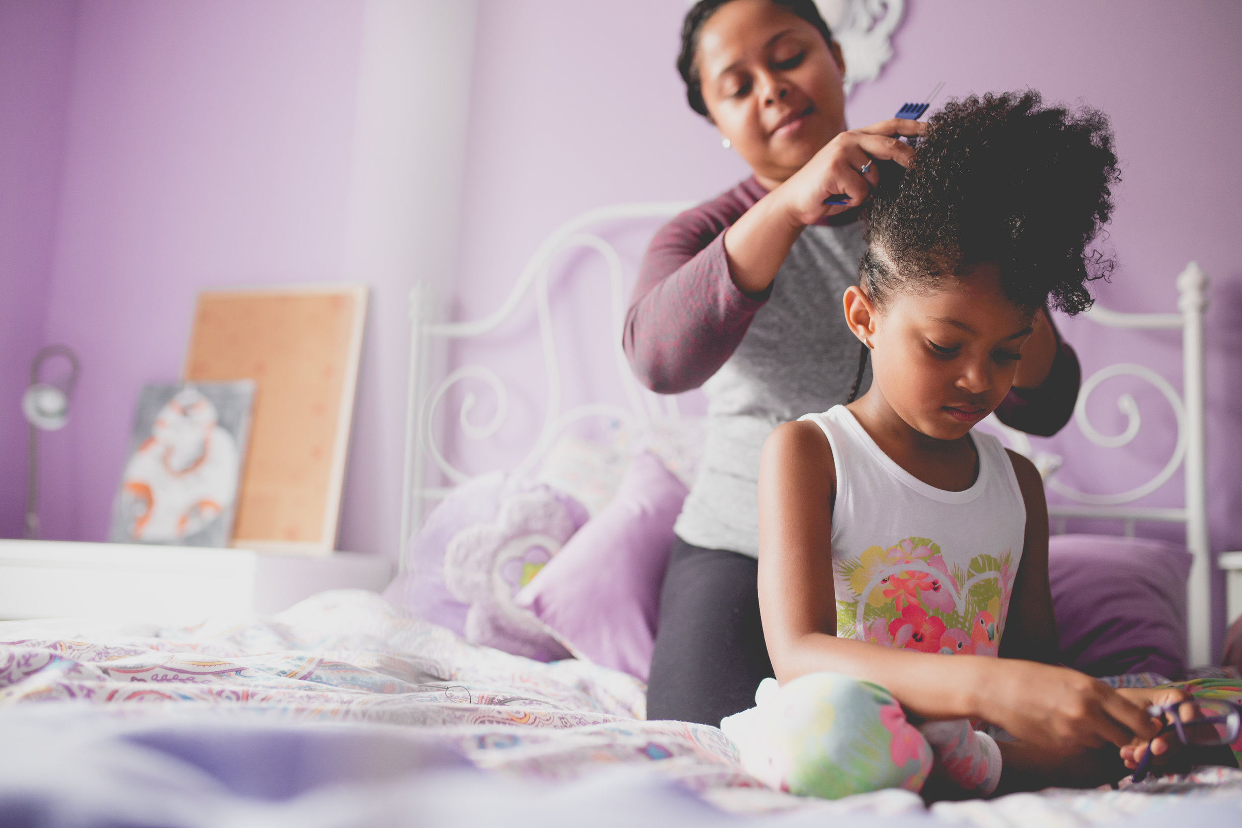 Mom and daughter on bed while mom puts cornrows in daughter's hair