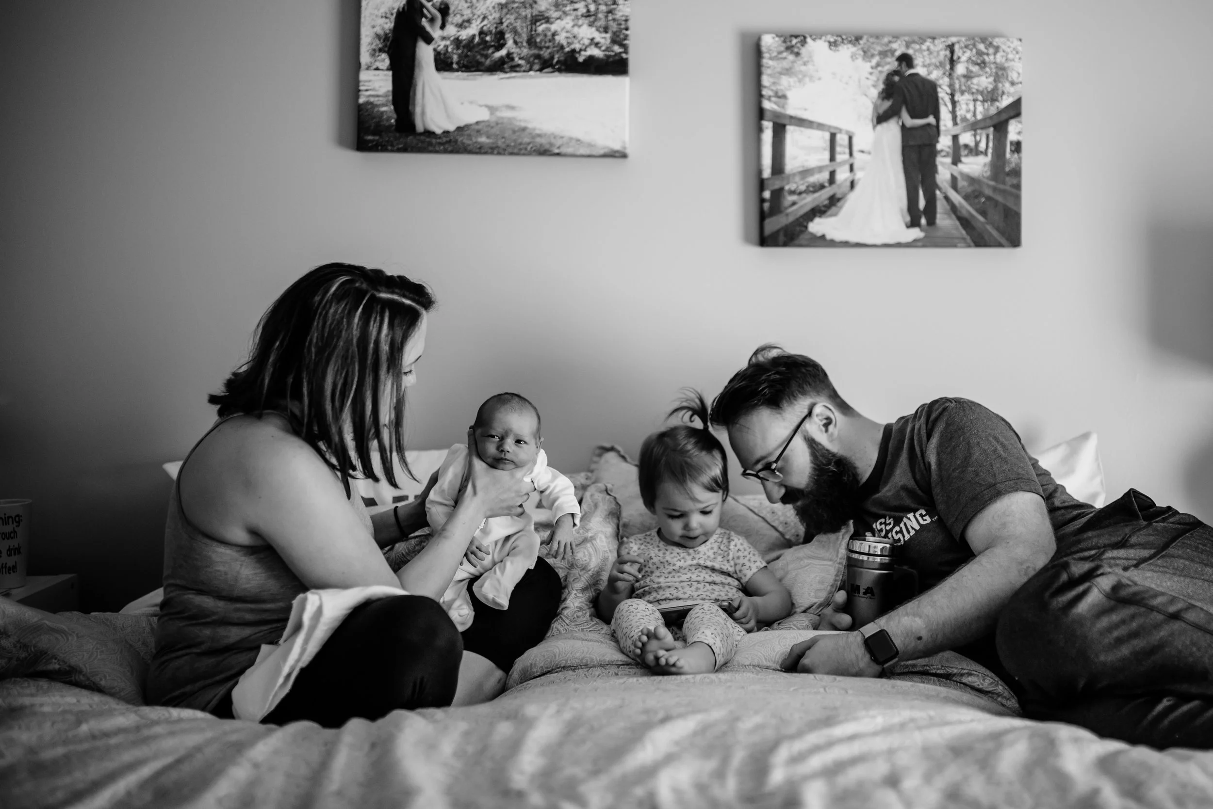 Family gathered on bed; Mom burps infant, Dad and older child look at phone
