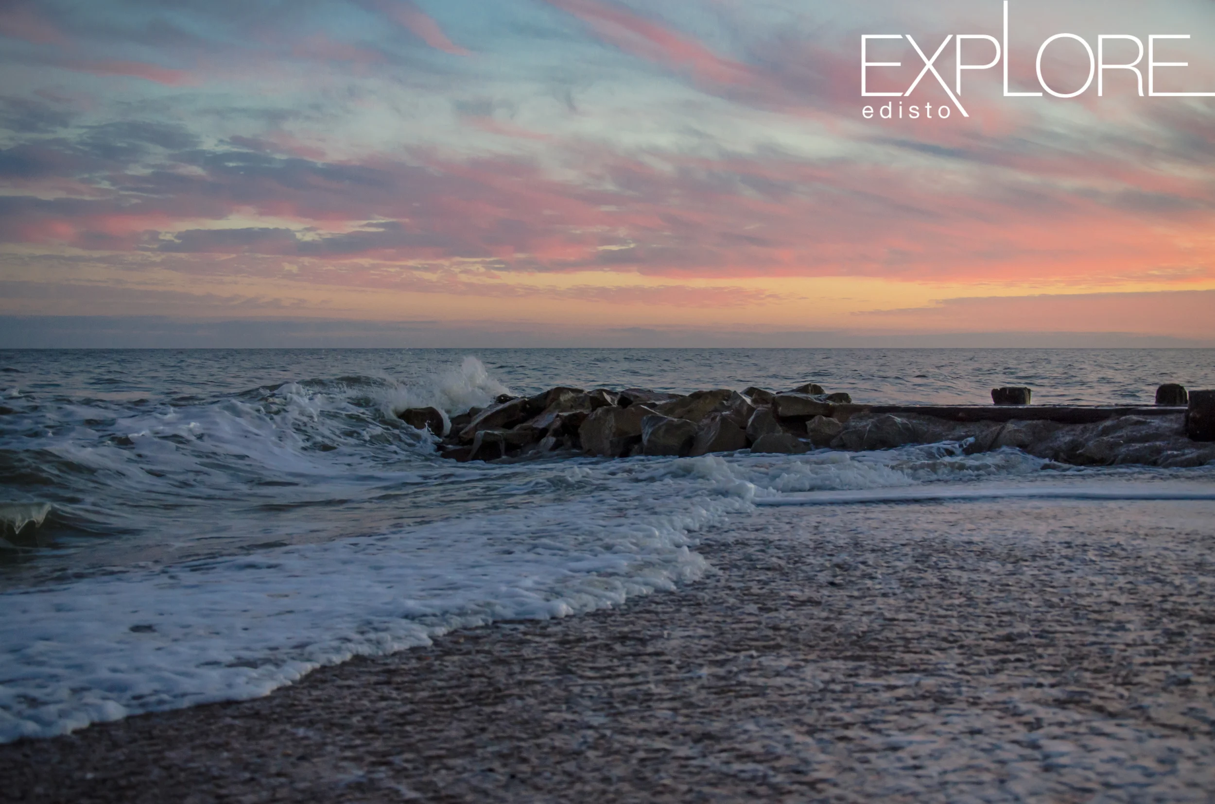 Pink, orange and blue sky over waves crashing on rocks.