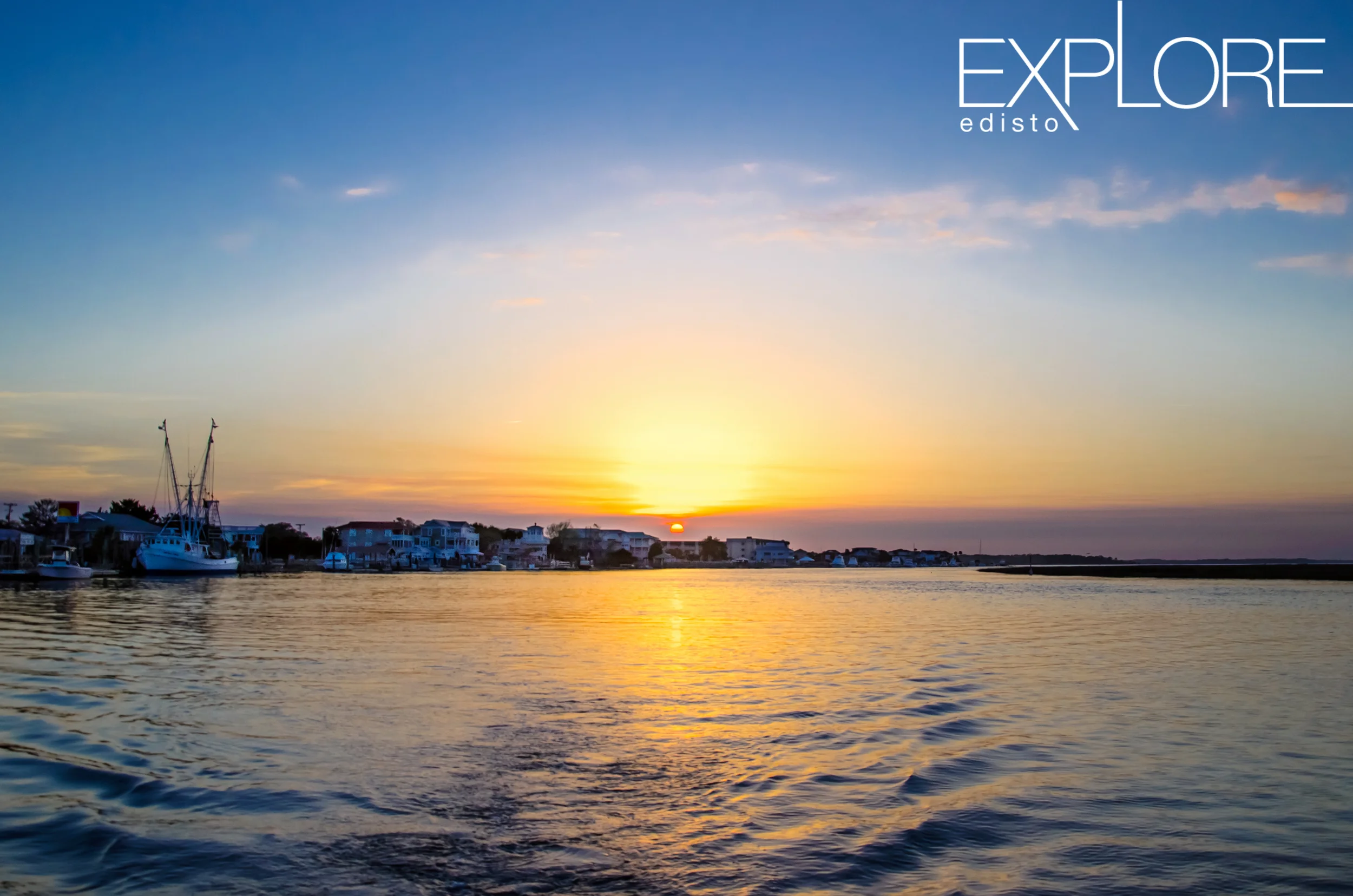 View of wake behind boat with shrimp boat and houses at sunset in the distance.